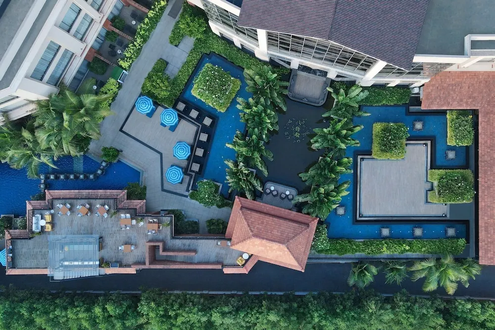 An aerial view shows a large landscaped pool courtyard with loungers, umbrellas, tropical plants and surrounding hotel buildings.