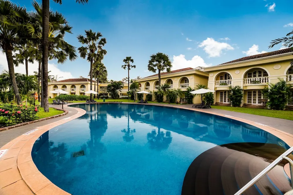 A large outdoor pool is surrounded by palm trees, loungers, umbrellas and colonial-style buildings.