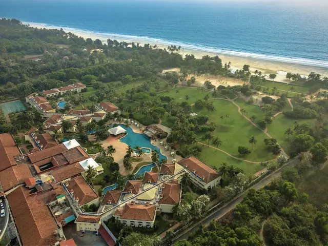 An aerial view shows a tropical resort, landscaped grounds, swimming pools, and a sandy beach beside the sea.