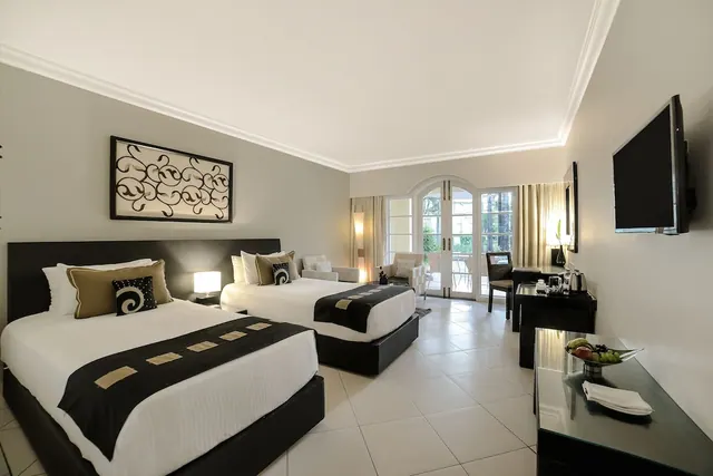 A bright hotel bedroom contains two twin beds, a seating area, a desk, and glass doors opening to a balcony.