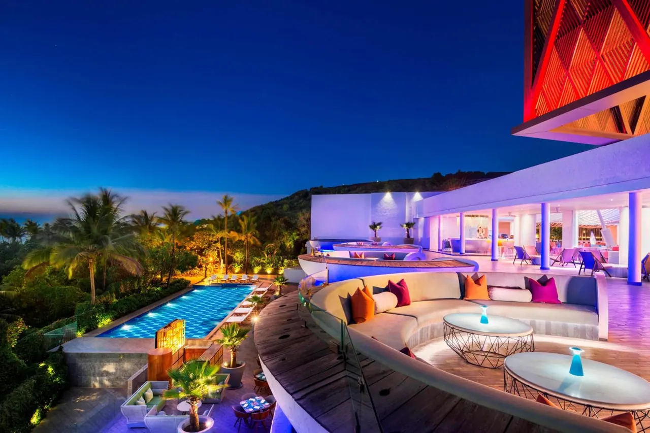 An illuminated pool and lounge terrace overlook tropical gardens and a wooded hillside.