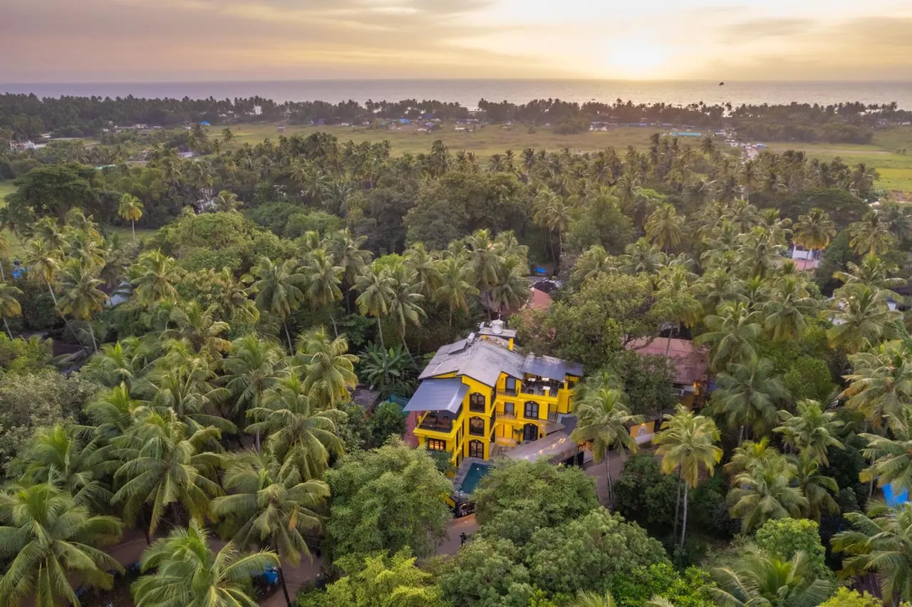 An aerial view shows a yellow hotel surrounded by coconut palms and tropical greenery with the sea beyond.
