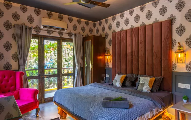 A furnished bedroom with a large bed, patterned walls, tall glass doors and greenery visible outside.