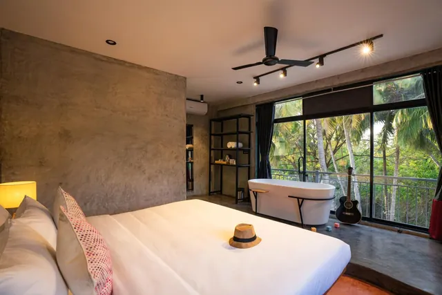 A bedroom with a large bed, freestanding bathtub, ceiling fan, and balcony overlooking palm trees.